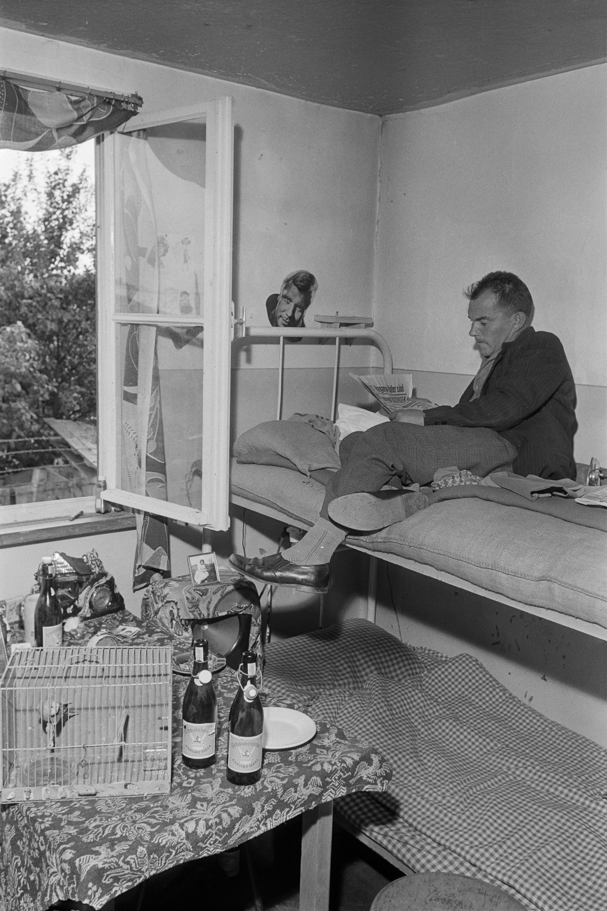 Zu sehen ist ein Mann, der auf einem Hochbett sitzt und liest. Daneben steht ein Tisch auf dem unter anderem Bierflaschen und ein Vogelkäfig stehen.