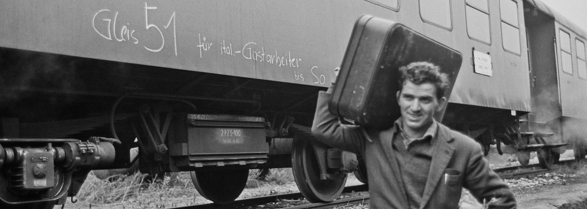 Zu sehen ist ein Mann der einen Koffer trägt. der Zug hintet ihm hat eine Aufschrift über italienische gastarbeiter.