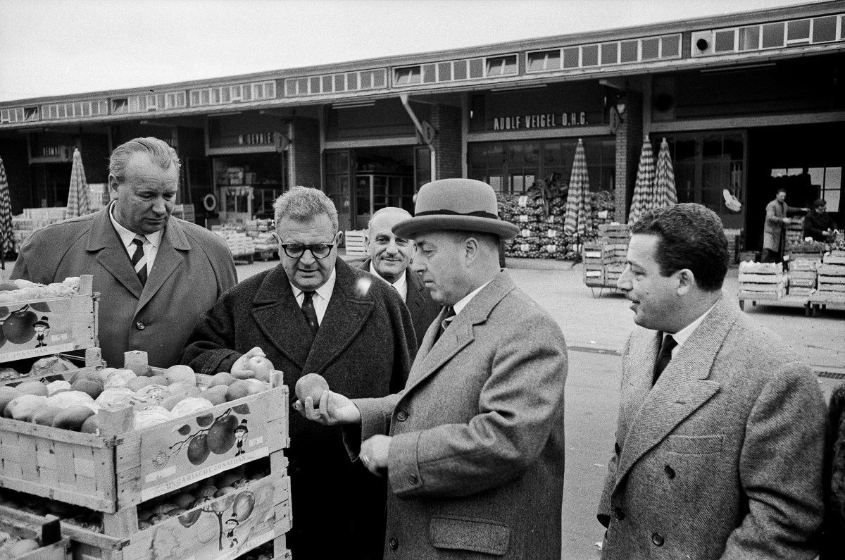 Zu sehen sind Männer,  die um Obstkisten herumstehen und das Obst begutachten.