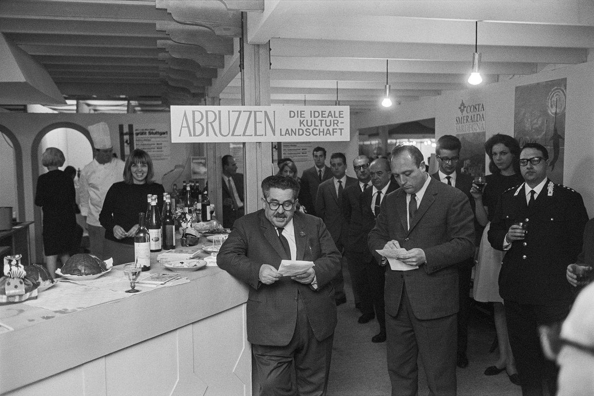 Zu sehen ist ein Mann der an eine Theke gelehnt ist und spricht während er von einem Zettel abliest. Um ihn herum stehen Menschen und hören ihm zu.