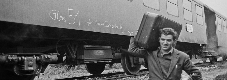 Zu sehen ist ein Mann der einen Koffer über Bahngleise trägt. Hinter ihm ist ein Zug der auf die Ankunft von italienischen Gastarbeitern hinweist.