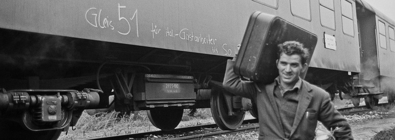 Zu sehen ist ein Mann der einen Koffer über Gleise trägt. Hinter ihm ist ein Waggon eines Zuges.