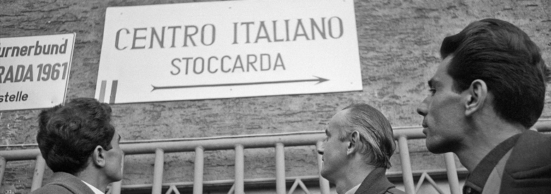 Zu sehen sind drei Männer, die auf ein Schild blicken, welches mit einem Pfeil und Schrift auf das italiensiche Zentrum Stuttgart hinweist.
