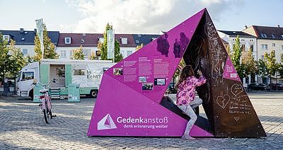 Ausschnitt aus Farbfotografie eines aufgestellten Vielecks in Pasewalk. Das Vieleck ist links rosa eingefärbt und mit Text- und Bildinformationen ausgestattet; rechts sind auf rostfarbenem Untergrund mit weißer Kreide Botschaften und Zeichnungen vorhanden. Ein Mädchen in rosa Jacke ist dabei, mit Kreide etwas hinzuzufügen. Im Hintergrund ein Fahrrad, Lastwagen und Häuser.