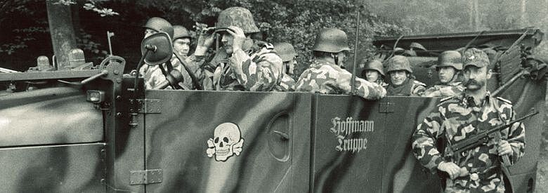 Ausschnitt aus dem Cover des Buchs Einzeltäter? Rechtsterroristische Akteure in der alten Bundesrepublik von Darius Muschiol. Schwarz-weiß-Fotografie der Wehrsportgruppe Hoffmann: In einem Auto sitzen mehrere in Camouflage gekleidete Männer mit Soldatenhelmen. Auf der vorderen linken Autotür ein Totenkopf; rechts davon auf einer weiteren Tür die Worte: Hoffmann Truppe. Rechts neben dem Auto stehend ein Mann mit Schnurrbart in Camouflage; eine Schildkappe tragend und in den Händen eine Schusswaffe haltend.