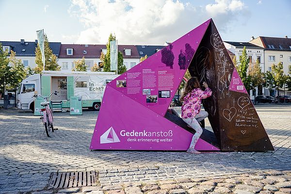 Farbfotografie eines aufgestellten Vielecks in Pasewalk. Das Vieleck ist links rosa eingefärbt und mit Text- und Bildinformationen ausgestattet; rechts sind auf rostfarbenem Untergrund mit weißer Kreide Botschaften und Zeichnungen vorhanden. Ein Mädchen in rosa Jacke ist dabei, mit Kreide etwas hinzuzufügen. Im Hintergrund ein Fahrrad, Lastwagen und Häuser.