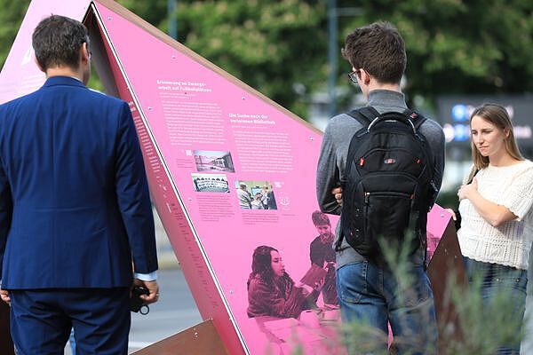 Farbfotografie eines aufgestellten Vielecks beim Deutschen Fußballmuseum Dortmund. Im Vordergrund zwei Personen, die mit dem Rücken zur Kamera das vor Ihnen stehende Vieleck betrachten, das auf rosa Hintergrund Text- und Bildinformationen zeigt (Überschriften: Erinnerung an Zwangsarbeit auf Fußballplätzen sowie Die Suche nach der verlorenen Bibliothek). Rechts im Hintergrund eine weitere Person, die das Vieleck betrachtet.