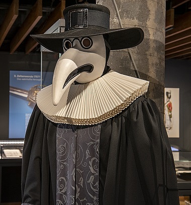 Das Bild zeigt einen Pestdoktor mit weißer Maske und schwarzem Umhang im Auststellungsraum.