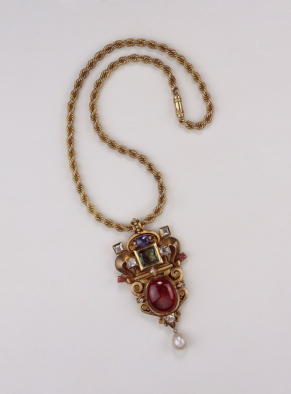 Zu sehen ist eine goldene Kette mit einem großen Anhänger. Dieser ist mit verschiedenfarbigen Edelsteinen verziert