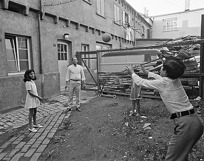 Zu sehen sind ein MAnn und drei Kinder, die in einem Hinterhof mit einem Ball spielen.