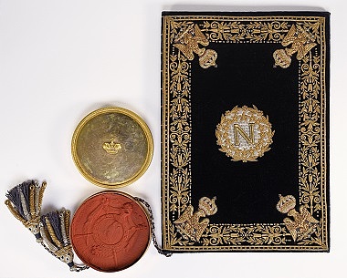 Zu sehen ist ein in Samt gebundener Vertrag, der mit goldenen Stickereien sowie einem goldenen N in der Mitte verziert ist. Angehängt eine ein großes Wachssiegel in einer goldenen Dose.