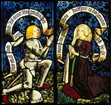 Zu sehen sind zwei Glasfenster. Links ist ein kniender Mann in Ritterrüstung und rechts eine kniende Frau in roter Robe. Sie sind einander zugewandt und beten dabei.